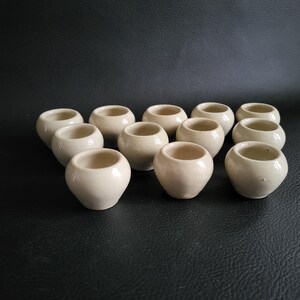 Peut inclure: Ensemble de petits pots en céramique blanc cassé disposés sur une surface sombre. Chaque pot présente un corps arrondi et une large ouverture. Les pots sont de taille et de forme similaires, suggérant un ensemble.
