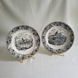 May include: Two black and white porcelain plates with decorative floral borders. The plates feature black and white illustrations of a horse and rider, with the text "Le Renard" and "Le Cerf" on each plate.