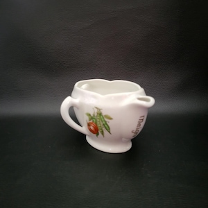Puede incluir: Una pequeña jarra de cerámica blanca con borde festoneado y dos asas. Está decorada con una colorida ilustración de verduras, incluyendo un tomate rojo y vainas de guisantes verdes. La palabra "Malign" está impresa en el lateral.