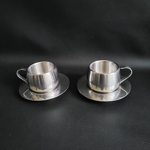 Peut inclure: Deux tasses à expresso en acier inoxydable avec leurs soucoupes assorties. Les tasses ont une forme arrondie avec de petites anses. Les soucoupes sont plates et circulaires. L'ensemble est en métal brillant et réfléchissant.