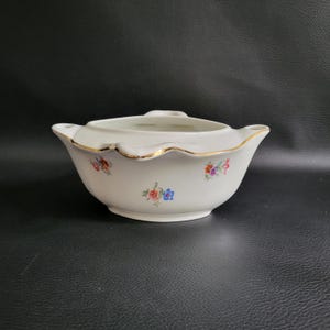 Puede incluir: Un cuenco de cerámica blanca con tapa y asas. El cuenco tiene un borde festoneado con ribete dorado y está decorado con pequeños grupos florales en rojo, azul y amarillo. El cuenco es probablemente vintage.