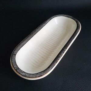 May include: An oval-shaped, white ceramic dish with a ridged interior. The rim is decorated with a black and silver dotted pattern. The dish is likely used for serving small portions of food or as a decorative item.