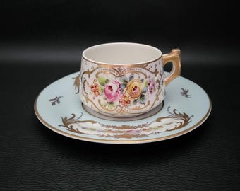 Może przedstawiać: Zestaw filiżanki i spodka do herbaty w stylu vintage. Filiżanka i spodek zdobione są kwiatowymi wzorami w kolorze różowym, żółtym i złotym. Spodek jest jasnoniebieski ze złotymi akcentami. Filiżanka ma złoty uchwyt.