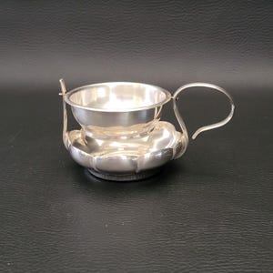 Könnte beinhalten: Ein kleines, silberfarbenes Tassen- und Untertassen-Set. Die Tasse hat einen Henkel und steht auf einer Untertasse mit gewelltem Rand. Das Set ist aus glänzendem Metall, das das Licht reflektiert. Die Tasse hat einen kleinen, befestigten Löffel.