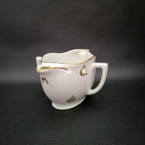 Puede incluir: Una cremera de cerámica blanca con ribetes dorados y detalles florales. La cremera tiene un asa y un pico vertedor, con la letra "M" dorada en el lateral. Sobre fondo negro.