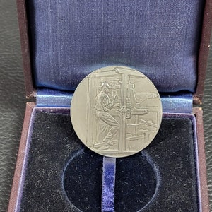 Peut inclure: Une médaille en argent avec une image en relief d'une personne utilisant une presse d'impression. La médaille est dans un coffret doublé de velours.