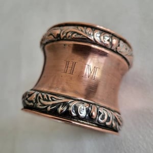 May include: A copper napkin ring with a silver floral design and the initials "H.M." engraved on the side.
