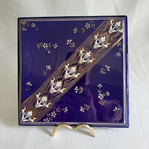 May include: Square, deep blue decorative tile featuring a diagonal band of white and gold ornate designs. Small white floral accents are scattered across the surface. The tile is displayed on a gold-coloured stand.