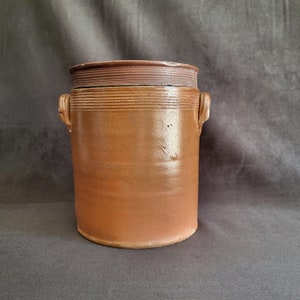 Puede incluir: Un tarro de cerámica marrón con tapa. El recipiente cilíndrico tiene dos asas pequeñas y estrías horizontales cerca de la parte superior. La tapa es de un tono marrón más oscuro. El tarro probablemente se utiliza para almacenar alimentos.