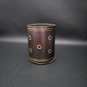Peut inclure: Un récipient cylindrique en cuir marron foncé avec des anneaux dorés. Le récipient a une bande dorée décorative en haut et en bas, et des anneaux dorés sont espacés uniformément sur le corps.