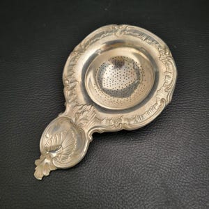 Könnte beinhalten: Ein silbernes Teesieb mit einem verzierten Griff und einer perforierten Schüssel. Das Sieb hat ein detailliertes, geprägtes Design um den Rand und den Griff und ist wahrscheinlich aus Sterlingsilber gefertigt. Das Teesieb ist für eine Teetasse konzipiert.