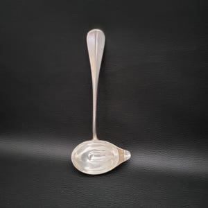 Puede incluir: Un cucharón de plata con un mango largo y delgado y un cuenco redondeado. El cuenco tiene un pequeño pico en un lado.