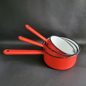 Puede incluir: Un juego de tres cacerolas rojas de esmalte con mangos largos. Las cacerolas están anidadas, con interiores blancos y ribetes negros. Los mangos son de un rojo brillante, y las ollas están dispuestas sobre una superficie oscura.