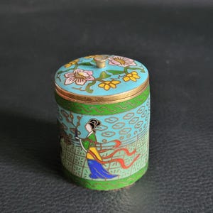 Può includere: Piccola scatola decorativa in cloisonné, rotonda e con coperchio. Il coperchio turchese presenta motivi floreali e un bottone dorato. I lati raffigurano una donna in abito tradizionale, su sfondo verde e blu. Rifiniture dorate.