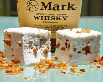 May include: Two white marshmallows topped with crispy brown bits, sitting on a light blue surface. A bottle of Maker's Mark Kentucky Straight Bourbon Whisky is visible in the background.