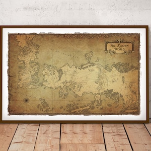 May include: A framed vintage-style map of "The Known World" with a brown frame and white border. The map features detailed landmasses in shades of brown and tan, with a grid overlay. The artwork is displayed on a wooden floor against a white wall.