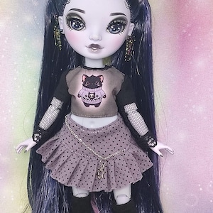 May include: A fashion doll with long dark purple hair, pale skin, and large grey eyes. The doll wears a short-sleeved grey crop top with a black cat graphic, a grey polka-dot pleated skirt with a silver chain, fishnet sleeves, and black boots.