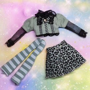 Può includere: Un completo per bambola composto da un top a maniche lunghe verde salvia con bordo in pizzo nero, un fiocco nero e maniche in rete, una gonna plissettata con stampa leopardata nera e grigia e un paio di calze al ginocchio a righe grigie e bianche.
