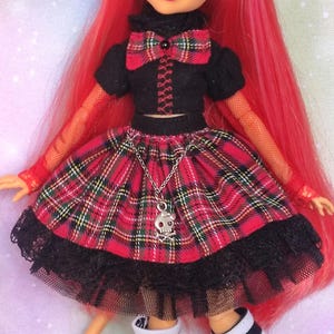 May include: A doll with long red hair wearing a black short-sleeved top with a red plaid bow, a matching red plaid skirt with a silver skull charm on a chain, black lace trim, orange fishnet sleeves, and black and white striped knee-high socks.
