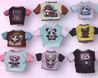 Könnte beinhalten: Eine Sammlung von neun Miniatur-Puppen-T-Shirts mit verschiedenen grafischen Drucken stilisierter Kreaturen, darunter Drachen, Fledermäuse und Katzen. Jedes T-Shirt hat kurze Ärmel und eine eigene Farbkombination, angeordnet in einem Raster.