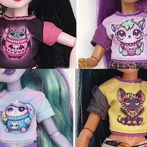 K&ouml;nnte beinhalten: Vier Puppenoberteile mit kurzen &Auml;rmeln. Jedes Shirt zeigt ein anderes Cartoon-Kreaturen-Design. Die Shirts haben verschiedene Farbkombinationen, darunter Schwarz und Rosa, Lila und Wei&szlig;, Blau und Pastell sowie Gelb und Grau. Die Oberteile sind Teil eines Puppenkleidungssets.