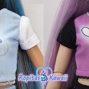 K&ouml;nnte beinhalten: Zwei Puppenoberteile: eines hellblau mit einem Care Bear auf einer Wolke, das andere lila mit einer Zeichentrickfigur im Fledermauskost&uuml;m. Beide Oberteile haben kurze &Auml;rmel und dekorative Verzierungen.