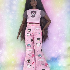K&ouml;nnte beinhalten: Puppe in einem rosa und grauen Crop-Top mit einem Cartoon-Motiv und einer rosa Hose mit Muster. Die Puppe hat lange dunkle Haare und schwarze Schuhe. Das Outfit ist ein Pyjama-Set.