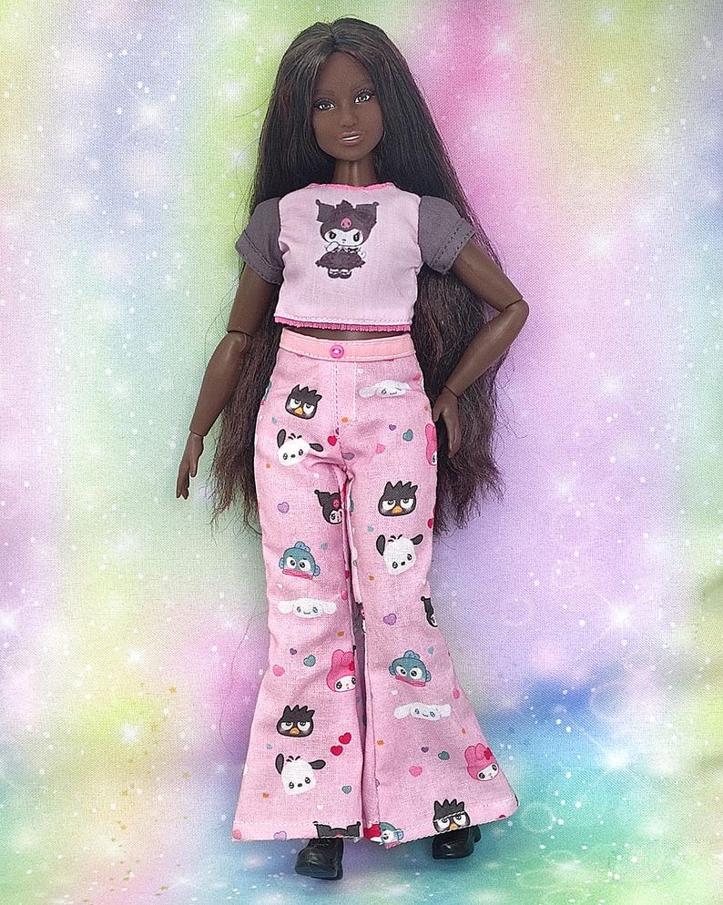K&ouml;nnte beinhalten: Puppe in einem rosa und grauen Crop-Top mit einem Cartoon-Motiv und einer rosa Hose mit Muster. Die Puppe hat lange dunkle Haare und schwarze Schuhe. Das Outfit ist ein Pyjama-Set.