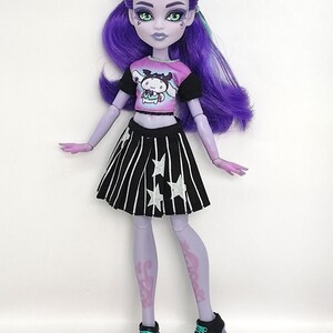 K&ouml;nnte beinhalten: Puppe mit violetter Haut, Haaren und Augen, tr&auml;gt ein rosa-schwarzes Oberteil mit einer Cartoonfigur, einen schwarz-wei&szlig; gestreiften Rock mit Sternen und schwarze Plateauschuhe. Die Puppe hat ein einzigartiges, stilisiertes Aussehen.