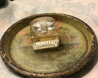 May include: A clear glass bottle of Chloe perfume on a green and brown metal tray with a faded floral design. The bottle has a square shape with a textured surface and the word "Chloe" is printed on the top.