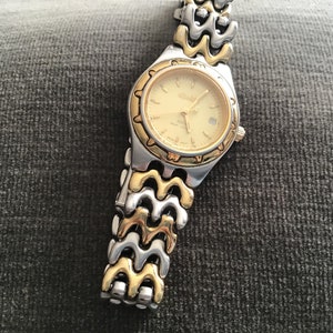 May include: A gold and silver two-tone watch with a gold-tone face and a date window. The watch has a wave-like band.