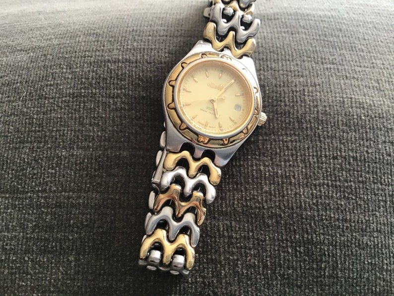 May include: A gold and silver two-tone watch with a gold-tone face and a date window. The watch has a wave-like band.
