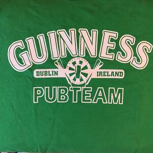 May include: Green t-shirt with the word "GUINNESS" in large white letters. Below the word "GUINNESS" is a white graphic of a dartboard with darts and the words "DUBLIN IRELAND". The words "PUBTEAM" are printed in white below the graphic.