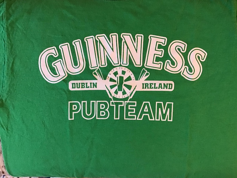 May include: Green t-shirt with the word "GUINNESS" in large white letters. Below the word "GUINNESS" is a white graphic of a dartboard with darts and the words "DUBLIN IRELAND". The words "PUBTEAM" are printed in white below the graphic.