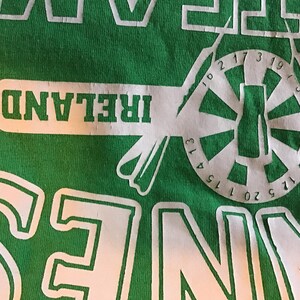 May include: Green t-shirt with white graphic design. The design includes the word "IRELAND" in a white rectangular box, and a circular design with numbers and a central shape resembling a bottle.