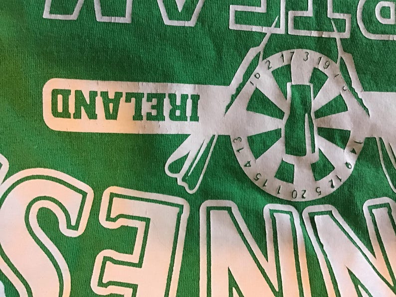 May include: Green t-shirt with white graphic design. The design includes the word "IRELAND" in a white rectangular box, and a circular design with numbers and a central shape resembling a bottle.