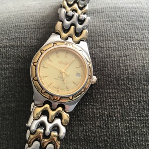 May include: A gold and silver two-tone watch with a gold-tone face and a date window. The watch has a gold-tone band with a wave-like design.