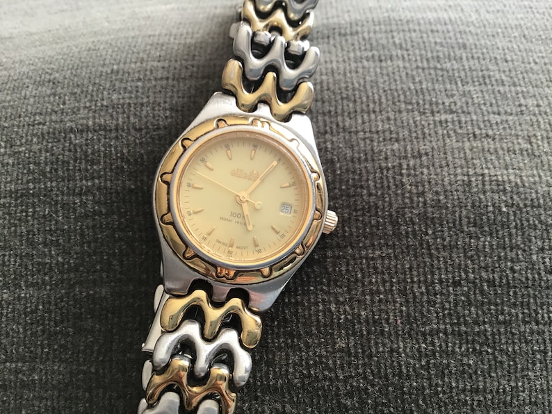 May include: A gold and silver two-tone watch with a gold-tone face and a date window. The watch has a gold-tone band with a wave-like design.
