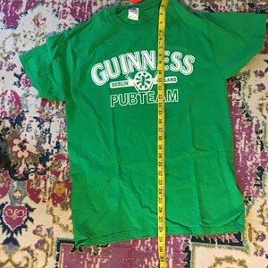 May include: Bright green t-shirt with the Guinness logo in white, including the words "Dublin" and "Land". The text "PUBTEAM" is below the logo. A yellow measuring tape is next to the shirt.