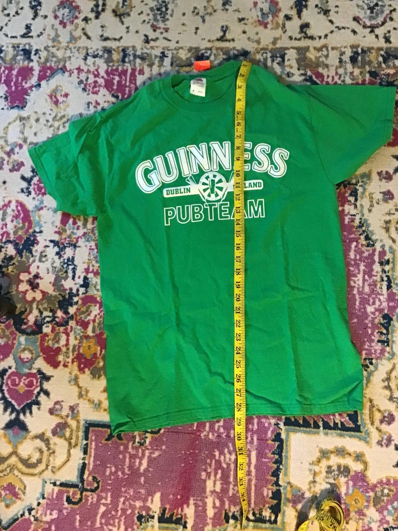 May include: Bright green t-shirt with the Guinness logo in white, including the words "Dublin" and "Land". The text "PUBTEAM" is below the logo. A yellow measuring tape is next to the shirt.