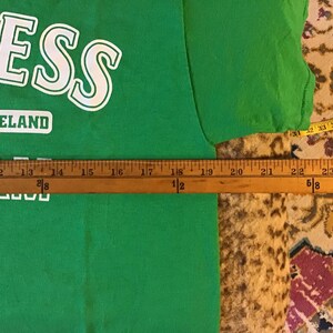 May include: A green t-shirt with the word "NESS" in white block letters. Below the word "NESS" is the word "IRELAND" in a white rectangle. A wooden ruler is placed across the shirt, measuring approximately 24 inches.