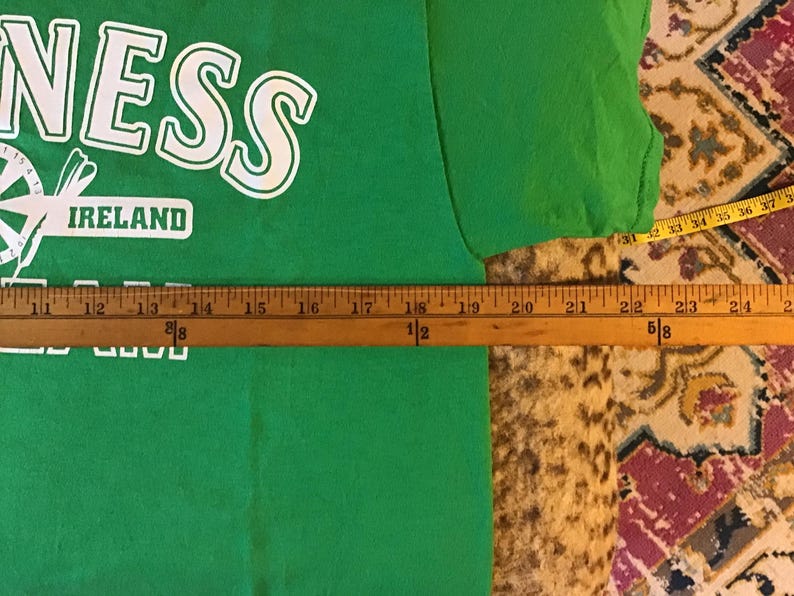 May include: A green t-shirt with the word "NESS" in white block letters. Below the word "NESS" is the word "IRELAND" in a white rectangle. A wooden ruler is placed across the shirt, measuring approximately 24 inches.