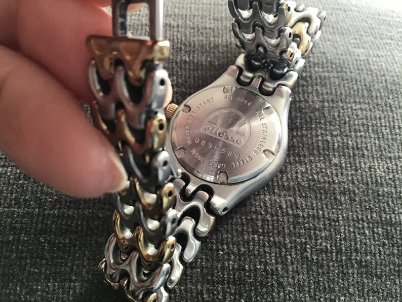 May include: A silver and gold watch with a stainless steel band. The watch face is obscured, but the back of the watch has the word "ellesse" printed on it.