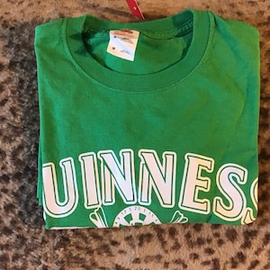 May include: A folded green t-shirt with the word "GUINNESS" in white block letters across the front. Below the text is a white graphic. The shirt has a crew neck and a tag attached to the collar.