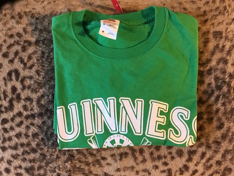 May include: A folded green t-shirt with the word "GUINNESS" in white block letters across the front. Below the text is a white graphic. The shirt has a crew neck and a tag attached to the collar.