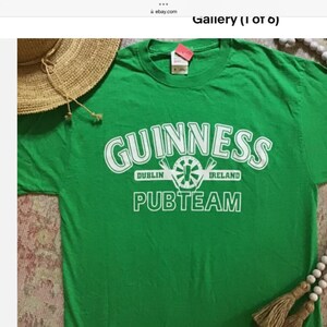May include: Green t-shirt with the Guinness logo in white, including the words "Dublin Ireland" and "Pubteam". A straw hat and beaded necklace with tassels are partially visible in the image.