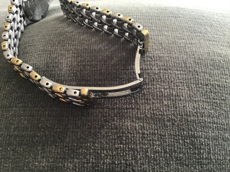 May include: A two-tone watch band with a silver and gold metal design. The band has a clasp closure.
