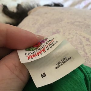 May include: A close-up of a white tag from a green t-shirt, held by a hand. The tag features the "Fruit of the Loom" logo and the words "Heavy HD." The tag also states "100% Cotton" and "Made in Honduras." The size "M" is printed on the tag.