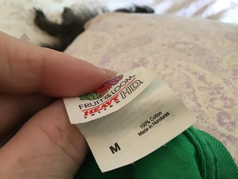 May include: A close-up of a white tag from a green t-shirt, held by a hand. The tag features the "Fruit of the Loom" logo and the words "Heavy HD." The tag also states "100% Cotton" and "Made in Honduras." The size "M" is printed on the tag.