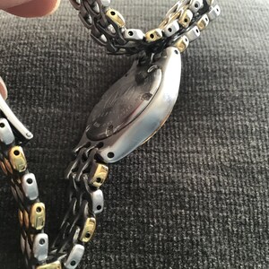 May include: A silver and gold two-tone watch with a stainless steel band. The watch has a round face and a silver case. The band is made of interlocking links.